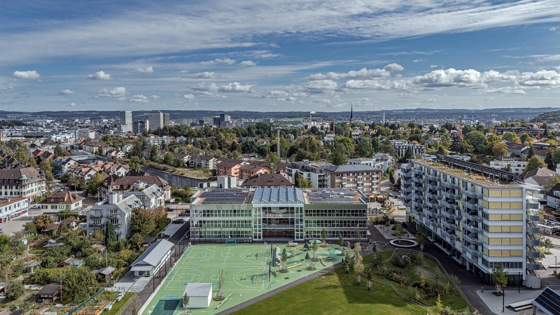 Luftaufnahme von Zürich mit Wohngebäuden und einem Schulkomplex mit grünen Sportfeldern und Spielplätzen im Vordergrund. Dahinter eine Mischung aus Bäumen, Wohnhäusern und modernen Hochhäusern unter teils bewölktem Himmel. Aufgenommen mit der DJI Mini 5 Pro. Aerial view of Zurich with residential buildings, a school complex with green sports fields and playgrounds in the foreground. In the background, trees, housing and modern high-rises under a partly cloudy sky. Captured with DJI Mini 5 Pro.
