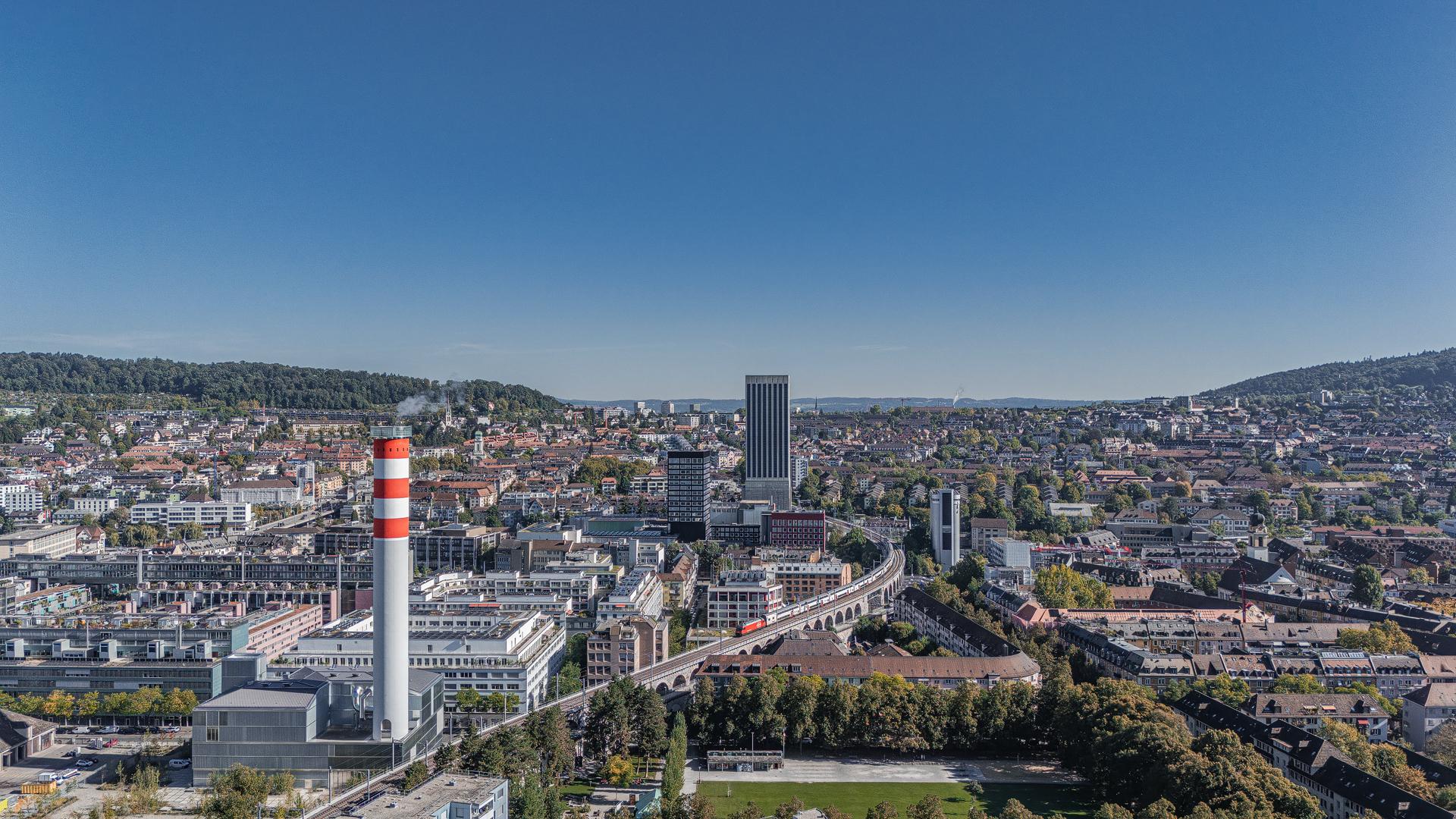 „Luftaufnahme von Zürich bei klarem Himmel. Im Vordergrund ein hoher Schornstein mit roten und weißen Streifen, daneben Industriegebäude. Dahinter die Stadt mit Wohnhäusern, modernen Hochhäusern und einem markanten Turm. Auf dem Viadukt fährt ein roter SBB-Zug. Im Hintergrund grüne Hügel.“ Arial view of Zurich on a clear day. In the foreground a tall red-and-white striped chimney, with residential areas, modern high-rises and a distinctive tower behind it. A red SBB train crosses the viaduct, with green hills in the distance.