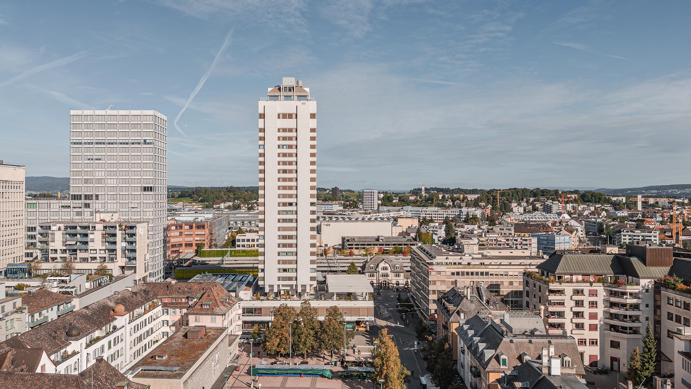 „Luftaufnahme von Zürich Oerlikon mit dem markanten Hochhaus ‚Oerlikon One‘ im Vordergrund. Davor die Bahngeleise und Bahnhofsanlagen, dahinter Wohn- und Geschäftshäuser sowie der grüne Hügelzug im Hintergrund unter leicht bewölktem Himmel. Aufgenommen mit der DJI Mini 5 Pro.“