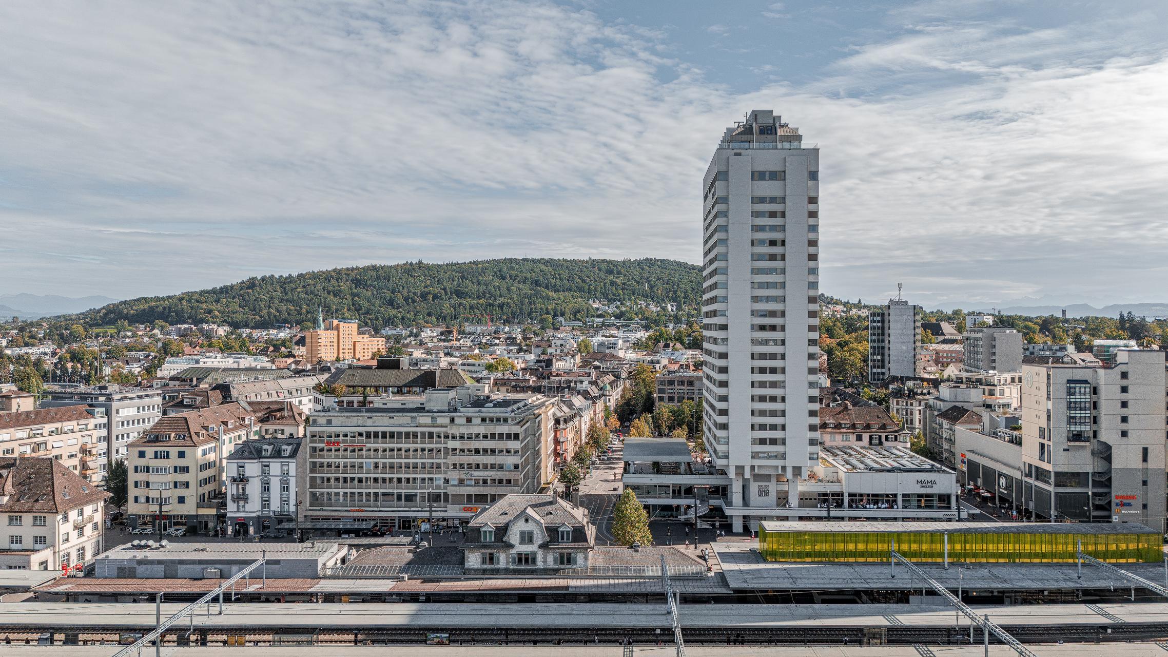 „Luftaufnahme von Zürich Oerlikon mit dem markanten Hochhaus ‚Oerlikon One‘ im Vordergrund. Davor die Bahngeleise und Bahnhofsanlagen, dahinter Wohn- und Geschäftshäuser sowie der grüne Hügelzug im Hintergrund unter leicht bewölktem Himmel. Aufgenommen mit der DJI Mini 5 Pro.“