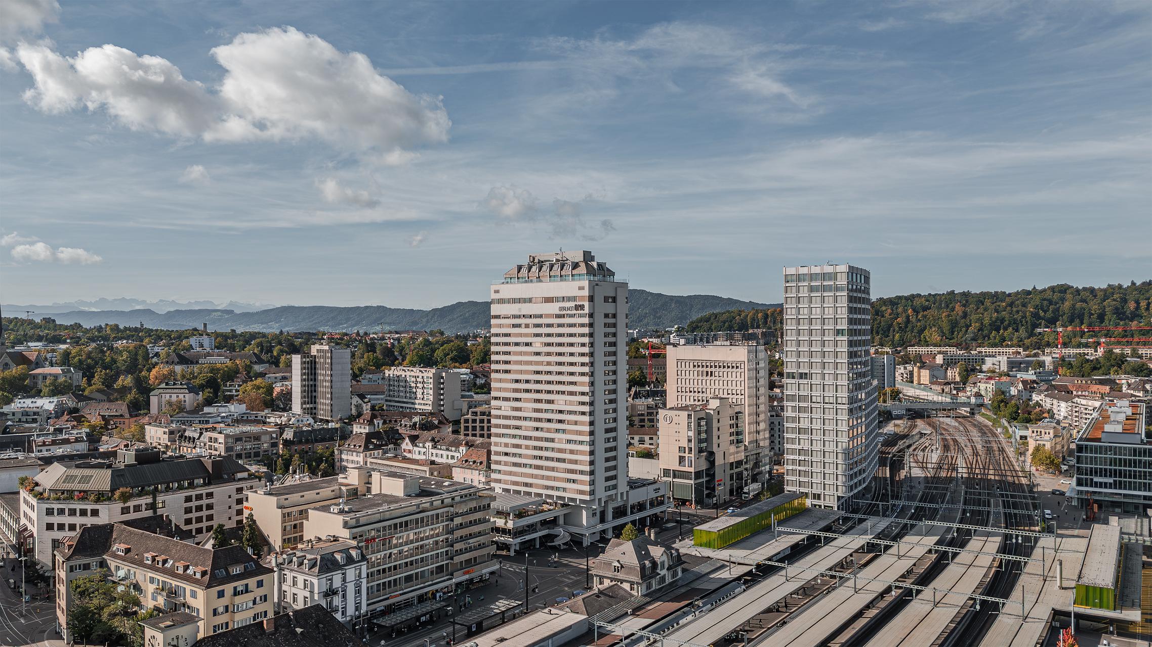 „Luftaufnahme von Zürich Oerlikon mit dem markanten Hochhaus ‚Oerlikon One‘ im Vordergrund. Davor die Bahngeleise und Bahnhofsanlagen, dahinter Wohn- und Geschäftshäuser sowie der grüne Hügelzug im Hintergrund unter leicht bewölktem Himmel. Aufgenommen mit der DJI Mini 5 Pro.“