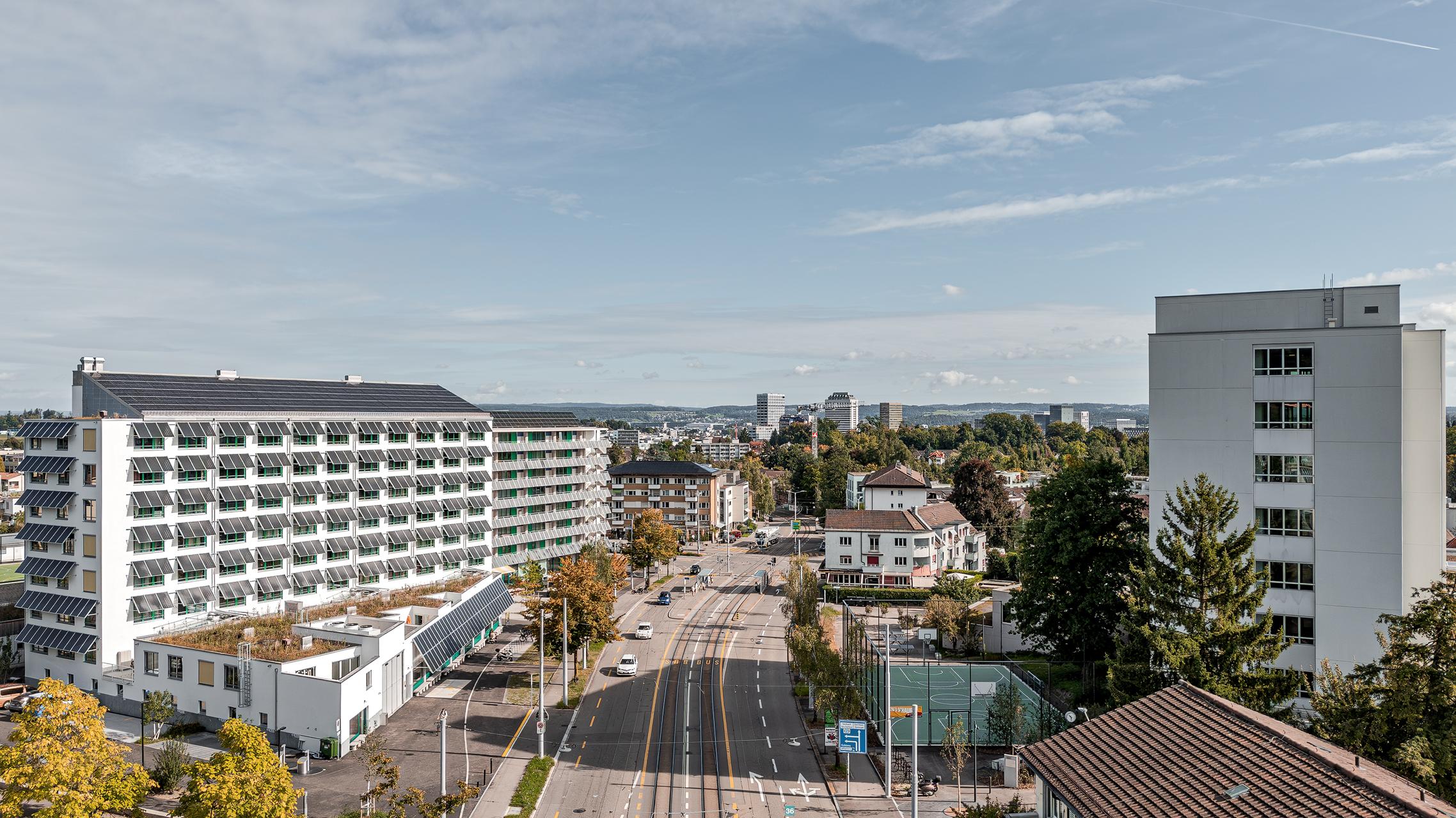 „Luftaufnahme von Zürich mit der Wohnüberbauung Guggach III im Vordergrund. Zu sehen sind moderne Wohngebäude und ein Schulkomplex mit Sport- und Spielplätzen. Im Hintergrund die Stadt mit Hochhäusern und grünen Baumreihen unter teils bewölktem Himmel. Entworfen von Donet Schäfer Reimer Architekten. Aufgenommen mit der DJI Mini 5 Pro.“
