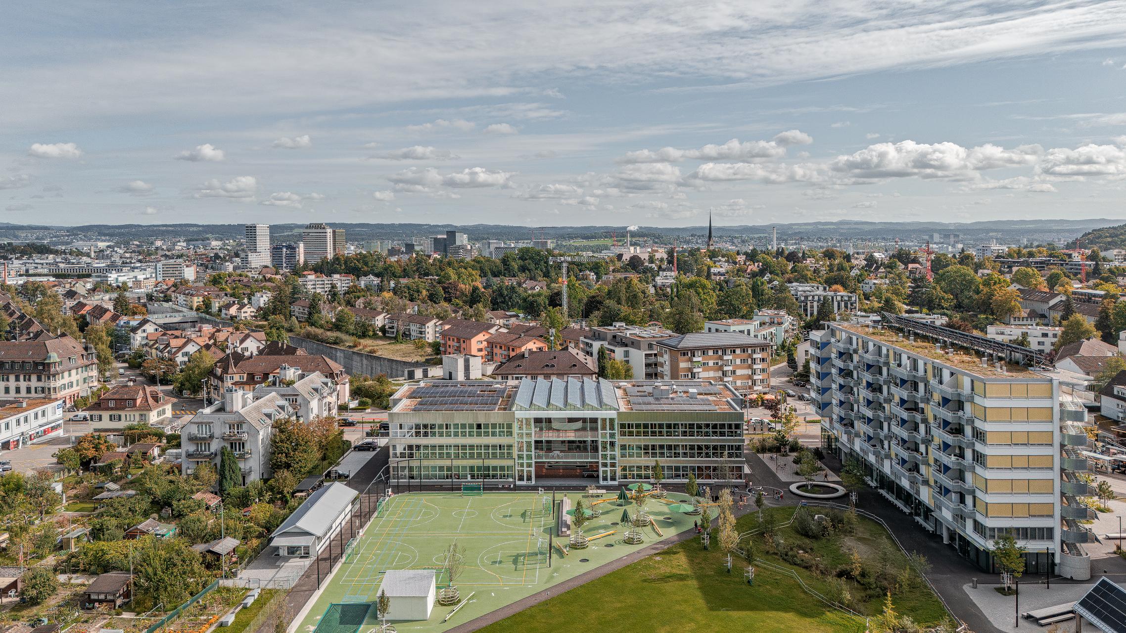 „Luftaufnahme von Zürich mit der Wohnüberbauung Guggach III im Vordergrund. Zu sehen sind moderne Wohngebäude und ein Schulkomplex mit Sport- und Spielplätzen. Im Hintergrund die Stadt mit Hochhäusern und grünen Baumreihen unter teils bewölktem Himmel. Entworfen von Donet Schäfer Reimer Architekten. Aufgenommen mit der DJI Mini 5 Pro.“